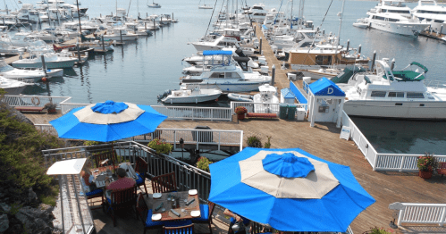 A marina with boats docked, featuring blue umbrellas over outdoor dining tables and a wooden walkway.