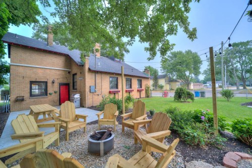 Cozy backyard with wooden chairs around a fire pit, leading to a brick house and green lawn under a cloudy sky.