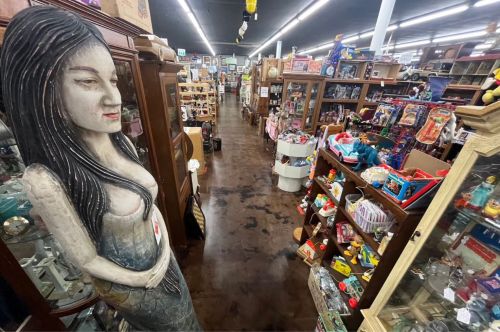A wide view of a cluttered antique store aisle, featuring various collectibles and a large wooden statue of a woman.