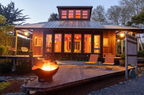 A cozy wooden house with large windows, a fire pit, and outdoor seating, illuminated at dusk.