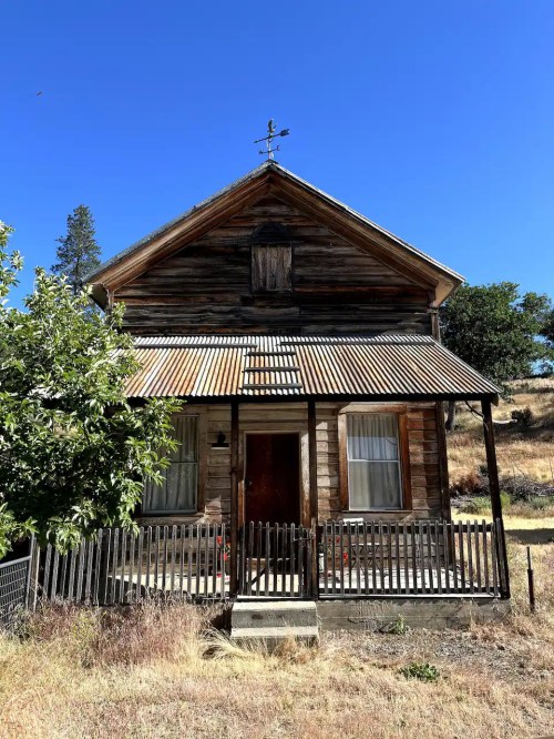 A rustic wooden house with a metal roof, surrounded by dry grass and trees, under a clear blue sky.