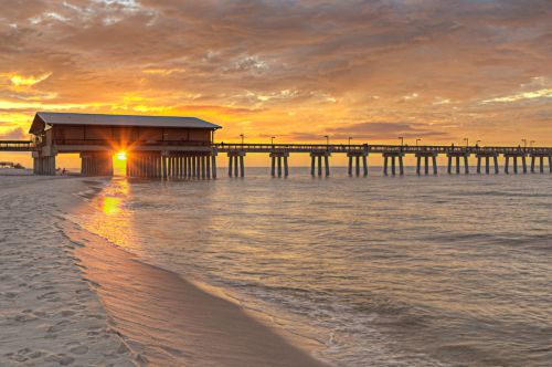 A pier extends over calm waters at sunset, with vibrant orange and yellow hues reflecting on the surface.