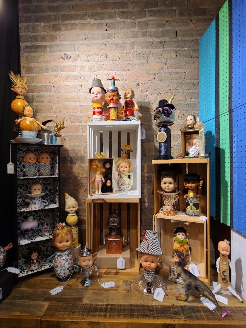 A display of vintage dolls and quirky figurines arranged on wooden crates against a brick wall.