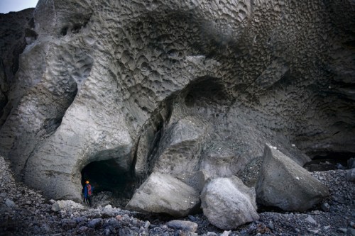 A person in a helmet stands near a large, textured rock formation with boulders on a rocky terrain.