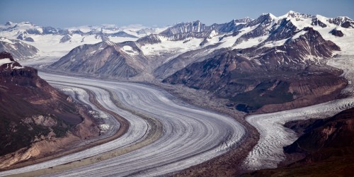 A sweeping view of a glacier winding through rugged mountains, with snow-capped peaks and a clear blue sky.