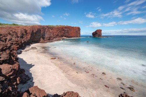 A serene beach with red cliffs, gentle waves, and a clear blue sky, creating a tranquil coastal scene.