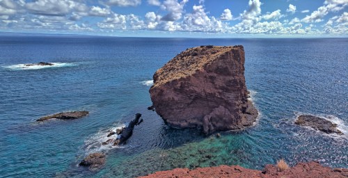 A rocky outcrop rises from clear blue waters, surrounded by small waves and a bright sky with scattered clouds.