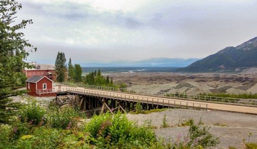 A red building on a wooden bridge overlooks a vast, rugged landscape with mountains in the distance and a hazy sky.