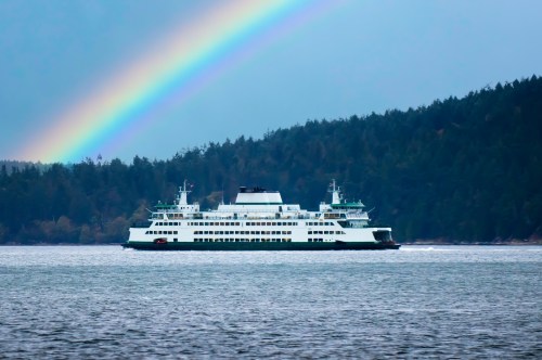 A ferry sails across calm waters beneath a vibrant rainbow and lush green hills in the background.