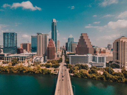 Aerial view of downtown Austin, Texas, featuring skyscrapers and a bridge over the river under a clear blue sky.
