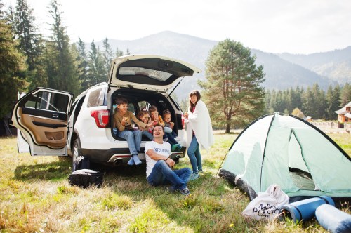 A family enjoys a picnic by their SUV, with a tent set up in a scenic forested area and mountains in the background.