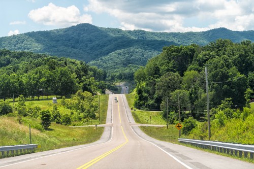 A winding road stretches through lush green hills under a partly cloudy sky.