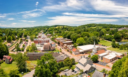 Aerial view of a small town with green hills, buildings, and a church under a bright blue sky with scattered clouds.