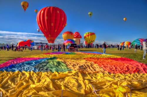 Colorful hot air balloons rise against a blue sky, with a vibrant rainbow fabric spread out on the ground below.