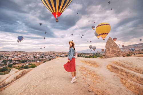 A woman in a red dress stands on a hill, surrounded by colorful hot air balloons in a scenic landscape.