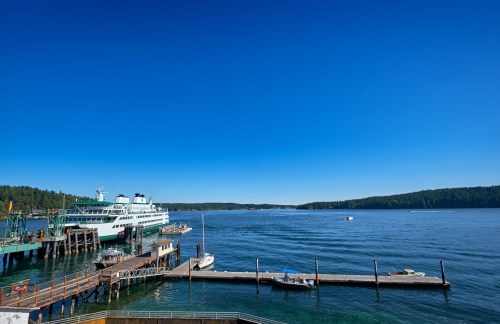 A ferry docked at a pier on a clear blue day, with calm waters and green trees in the background.
