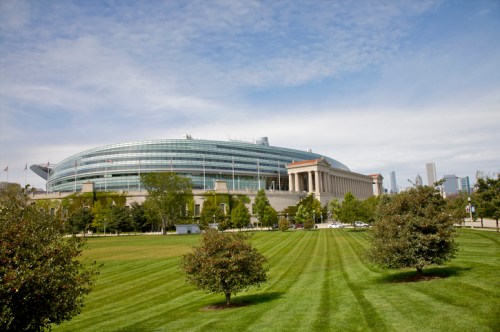 A wide view of a modern stadium surrounded by green grass and trees under a blue sky with scattered clouds.