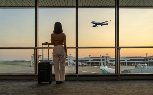 A traveler with a suitcase gazes out at an airplane taking off from an airport during sunset.