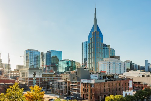 City skyline featuring modern buildings and a prominent spire, with clear blue skies and greenery in the foreground.