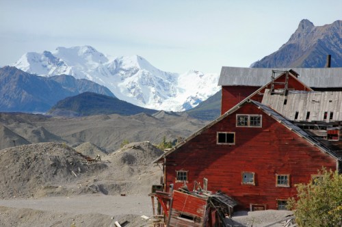 A weathered red building in a barren landscape, with snow-capped mountains in the background under a clear sky.