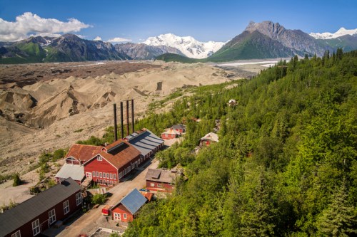 A scenic view of mountains, a glacier, and a historic red building surrounded by lush greenery and sandy terrain.
