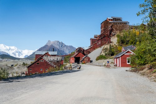 Historic red buildings on a gravel road, set against a backdrop of mountains and clear blue sky.