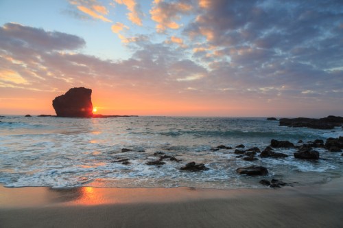 Sunset over the ocean with a rocky outcrop, clouds reflecting warm colors in the sky and gentle waves on the shore.