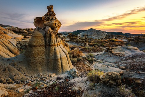 A unique rock formation stands against a colorful sunset in a rugged, arid landscape.