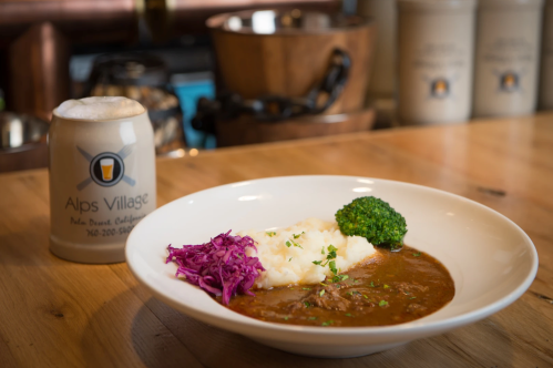 A bowl of stew with mashed potatoes, broccoli, and red cabbage, alongside a beer mug on a wooden table.