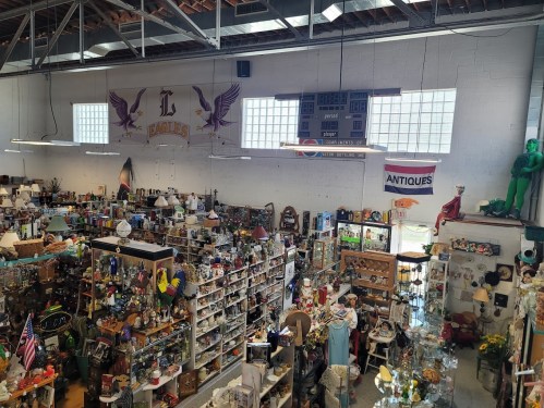 A spacious antique store filled with various collectibles, lamps, and decor, featuring high ceilings and large windows.