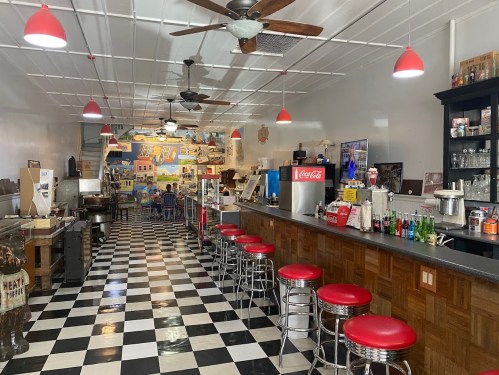 A retro diner interior with checkered floors, red stools, and a soda fountain, featuring colorful wall art.