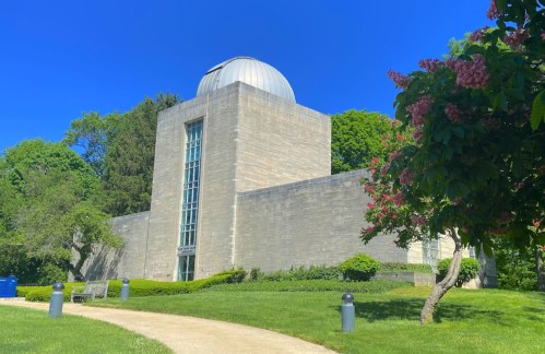 A modern stone observatory with a dome, surrounded by green trees and blooming flowers under a clear blue sky.