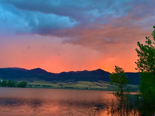 A serene lake at sunset, reflecting vibrant orange and purple hues against distant mountains and a cloudy sky.