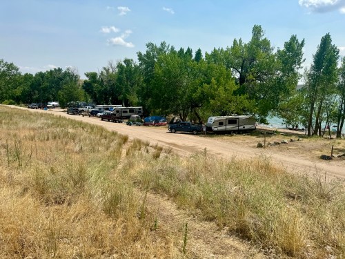 A gravel road lined with parked RVs and cars near a lake, surrounded by trees and grassy areas under a blue sky.