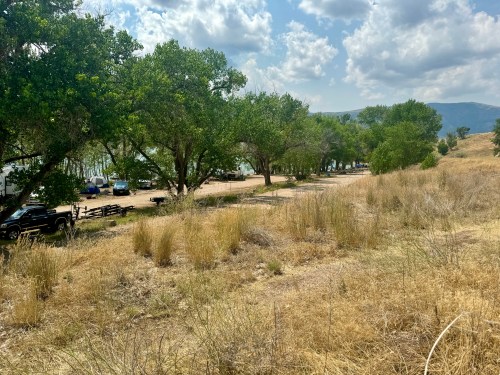 A scenic view of a grassy area with trees, parked cars, and a distant landscape under a partly cloudy sky.