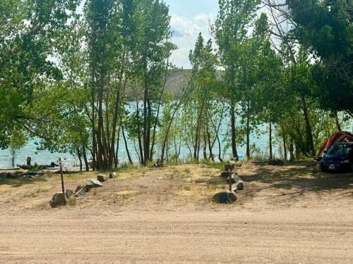 A serene lakeside scene with trees, a sandy area, and a few people relaxing by the water under a clear sky.