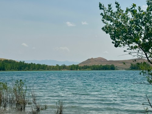 A serene lake surrounded by greenery and distant hills under a hazy sky.