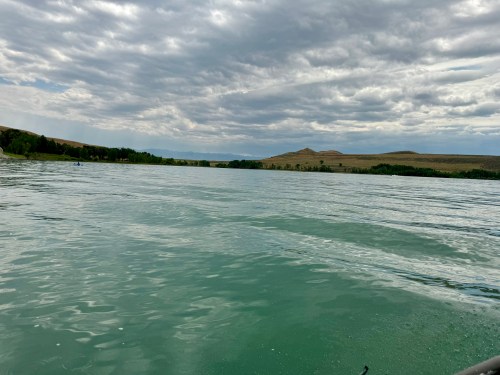 A calm lake reflects cloudy skies, surrounded by green trees and distant hills under a gray atmosphere.