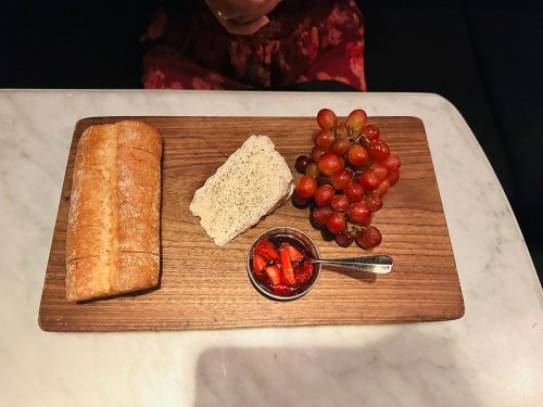 A wooden board with a loaf of bread, a slice of cheese, grapes, and a small dish of strawberry jam.