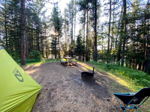 A serene campsite in a forest, featuring a tent, picnic table, fire pit, and surrounding trees.