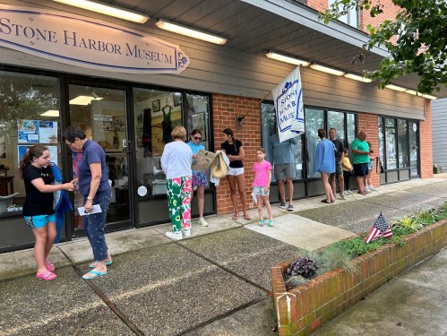 A line of people waiting outside the Stone Harbor Museum on a rainy day, with an American flag in the foreground.