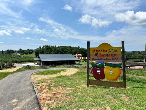 A colorful sign for Justus Orchard, with a path leading to the orchard and a building in the background under a blue sky.