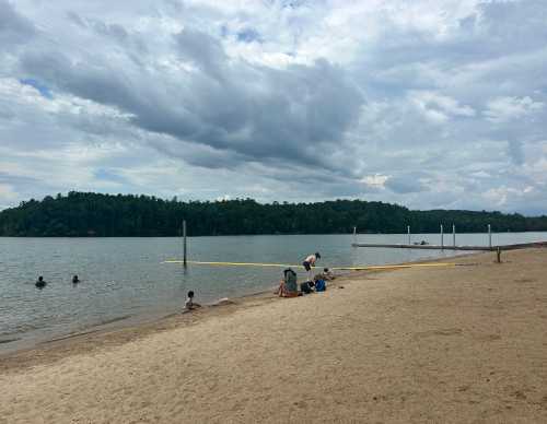 A sandy beach by a lake with people swimming and relaxing, surrounded by trees and cloudy skies.