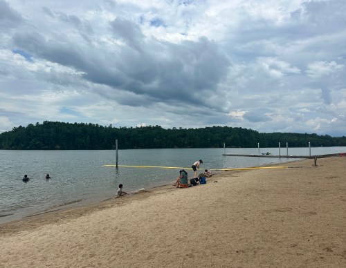 A sandy beach by a lake with people swimming and relaxing, surrounded by trees and cloudy skies.