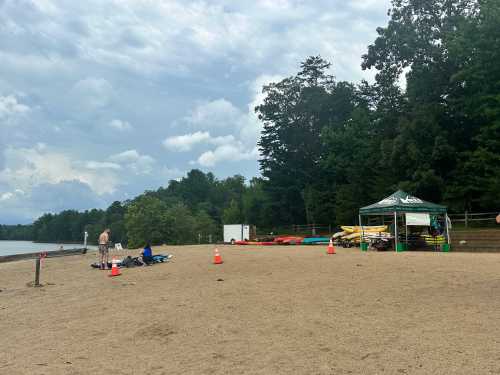 A sandy beach area by a lake with kayaks, a tent, and people preparing for water activities under a cloudy sky.