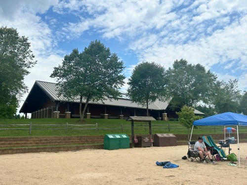 A sandy beach area with a pavilion in the background, trees nearby, and a person sitting under a blue canopy.