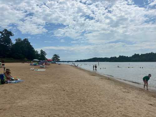 A sandy beach with people relaxing, swimming, and umbrellas under a partly cloudy sky by a calm lake.