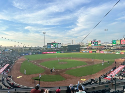A panoramic view of a baseball field with players on the field and spectators in the stands under a blue sky.
