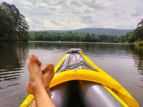 A person relaxes in a yellow kayak on a calm lake, surrounded by trees and mountains under a cloudy sky.