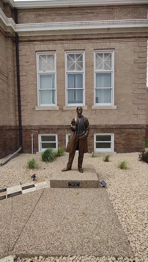 A bronze statue of a man in a suit stands outside a brick building, surrounded by gravel and small plants.